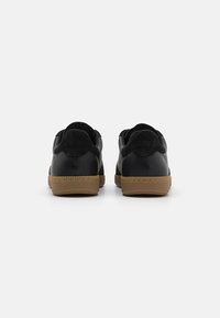 Adidași negri cu un exterior din piele netedă, accente din suede la călcâi și o talpă din cauciuc maro deschis, cu detalii texturate.