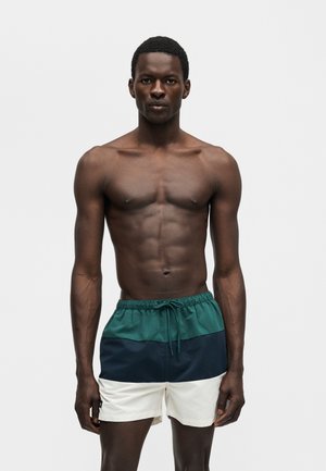 Αθλητικός άνδρας με σκουρόχρωμο δέρμα στέκεται, φορώντας πολύχρωμα μαγιό με μπλοκ χρωμάτων σε πράσινο, ναυτικό μπλε και λευκό, σε απλό φωτεινό φόντο.