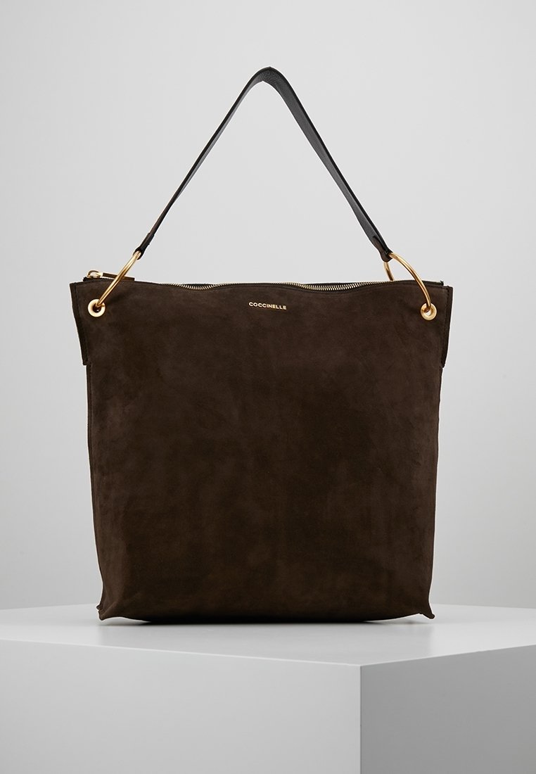 Sac fourre-tout en daim marron foncé avec des poignées en anneau doré et une bandoulière noire, posé sur une plateforme blanche.