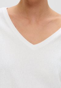Bílý top s výstřihem do V vyrobený z texturované látky s jemnými vertikálními pruhy. Nabízí volný střih a minimalistický design.