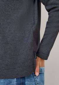 Camicia a maniche lunghe con righe scure, dal fit rilassato, con un'etichetta del marchio visibile e un tessuto texturizzato, abbinata a jeans azzurri chiari.