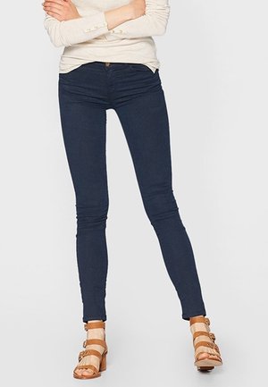 Jeans skinny bleu marine avec une texture lisse, dotés d'une fermeture à un bouton et de poches frontales, associés à des sandales à lanières tan.