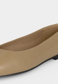 Μπεζ επίπεδο παπούτσι με χαμηλή μαύρη σόλα, στρογγυλή μύτη και λείο δερμάτινο επάνω μέρος ορατό από πλάγια γωνία.