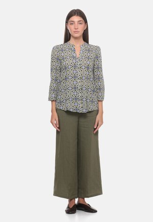 Donna in piedi che indossa una camicetta a maniche lunghe con fantasia e scollo a V e pantaloni larghi color oliva con scarpe scure senza lacci.