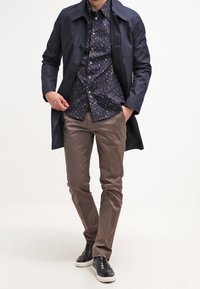 Námornická trench coat přes vzorovanou tmavou košili, spárovaná se světle hnědými kalhotami a černými teniskami. Oblečení má dob�ře padnoucí střih.