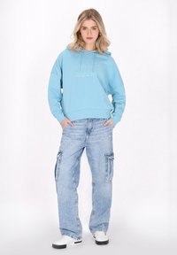 Sudadera recortada de color azul claro con un logotipo en el frente, combinada con unos jeans cargo de ajuste holgado en azul claro con bolsillos laterales y zapatillas blancas.