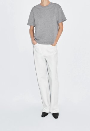 Osoba ubrana w szarą koszulkę z krótkim rękawem, wpuszczoną do białych, wysokich spodni z rękami w kieszeniach i czarne, spiczaste buty.