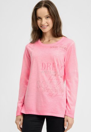 Woman with long brown hair wearing a bright pink long-sleeve shirt with mixed text designs and black pants, smiling slightly.