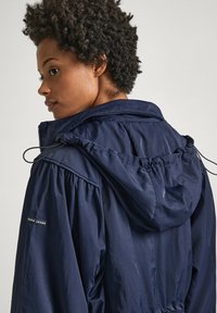 Veste légère bleu marine avec capuche à cordon de serrage, épaules froncées et un petit logo sur la manche. Texture lisse, design décontracté.