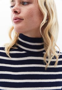 Navy och vit randig stickad tröja med hög krage, med ribbad textur och en figurnära design. Långt, böljande blont hår delvis synligt.