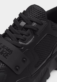 Μαύρο αθλητικό παπούτσι με πλέγμα και συνθετικά πάνελ, γλωττίδα από υφασμάτινο υλικό με υφή, στρογγυλά μαύρα κορδόνια και λουράκι με εμπορικό σήμα στο μπροστινό μέρος.