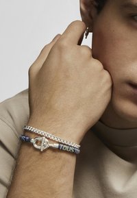 Pulsera de cadena de plata combinada con una pulsera tejida azul que presenta un cierre metálico, llevada en una mano sobre un fondo neutro.
