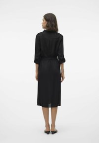 Abito nero a lunghezza ginocchio con colletto, maniche lunghe arrotolate fino al gomito e una cintura in tessuto. Texture liscia e design minimale.