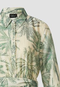 Chemise à manches longues boutonnée avec imprimé de feuilles de palmier vertes sur tissu beige, avec col et ceinture en tissu à la taille.