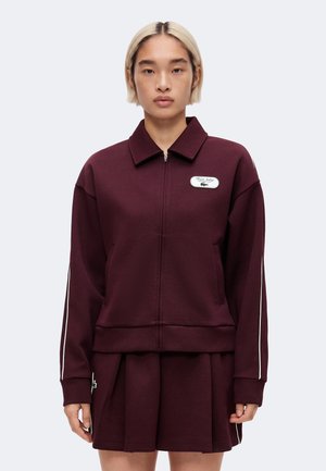 Persona con cabello rubio corto lleva una chaqueta burdeos con cremallera y una falda plisada a juego con un pequeño logo en el pecho y rayas en las mangas.