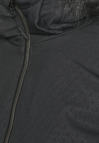 Primer plano de una chaqueta negra con cremallera visible y tejido con un sutil patrón geométrico, con capucha adornada con pelo.