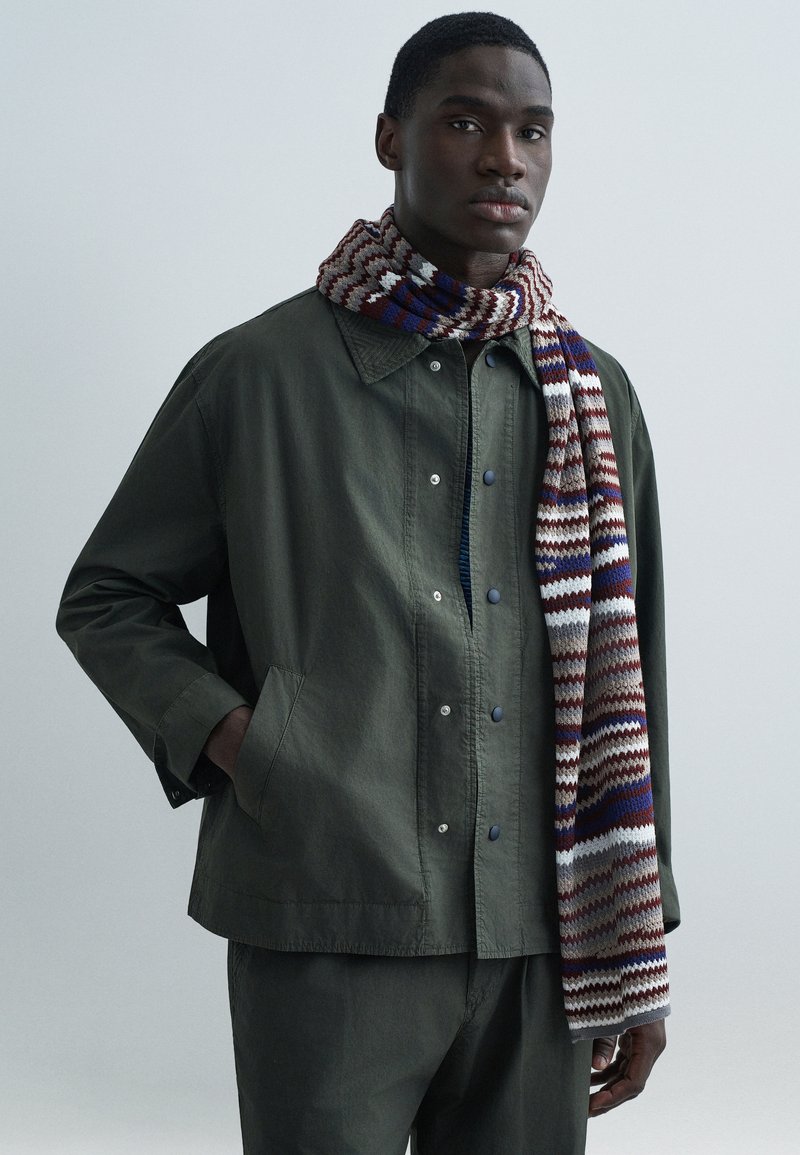 Olivovo zelená bunda so zapínaním na patentky, doplnená štvorfarebnou šatkou s pruhmi v červenej, modrej a bielej farbe.