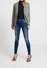 Kapeat siniset farkut, joissa hienovaraiset kulutetut yksityiskohdat, yhdistetty ruudulliseen bleiseriin, jossa on harmaan ja keltaisen sävyjä, valkoisella taustalla.