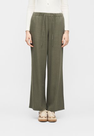 Persona che indossa pantaloni larghi di colore verde oliva con vita regolabile e scarpe da ginnastica beige con accenti maculati, in piedi contro uno sfondo chiaro e uniforme.