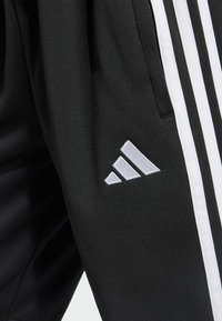 Zwarte sportbroek met witte zijstrepen en een witte geborduurde logo met drie diagonale staven. Glad stofoppervlak.