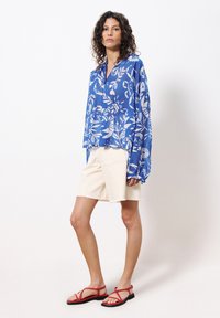 Bluse mit langen Ärmeln in Blau mit weißen Mustern, kombiniert mit cremefarbenen Shorts und roten Sandalen mit Riemen. Lockere Passform, leichter Stoff.
