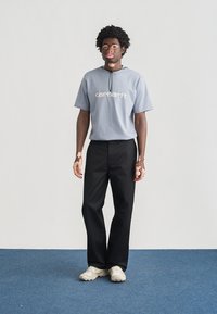 Γκρι κοντομάνικη μπλούζα με το λογότυπο "Carhartt" σε κρεμ απόχρωση, συνδυασμένη με μαύρο παντελόνι ίσιας γραμμής και κρεμ αθλητικά παπούτσια. Απλός, casual σχεδιασμός.