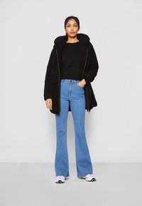 Haut à capuche en peluche noire avec fermeture éclair, porté sur un haut noir, assorti avec un jean évasé bleu et des baskets blanches. Tenue simple et décontractée.
