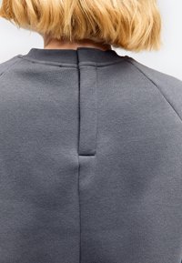 Bagfraudsigt af en person med kort blondt hår iført en grå sweatshirt med en lodret søm og stofflig ved halsen.