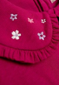 Fuksiaroosa topp, millel on volditud kaelus ja millel on roosa, valge ja lilla brodeeritud lilledega ornament. Pehme tekstuur, mille pind on veidi sooniline.