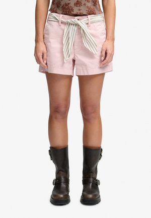Persoon draagt roze short met een gestrikte gestreepte stoffen riem en donkerbruine mid-kuit buckle laarzen, staat met armen ontspannen langs het lichaam.