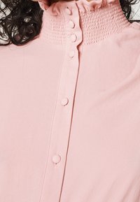 Blouse rose avec un col smocké haut, un bouton sur le devant et une texture lisse. Le tissu semble léger et doux.