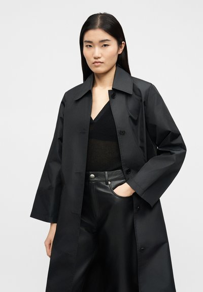 Γυναίκα που φοράει μαύρο μακρύ παλτό με κουμπιά, διάφανο μαύρο τοπ με V-λαιμόκοψη και μαύρο δερμάτινο παντελόνι, με το ένα χέρι στην τσέπη, σε απλό φόντο.