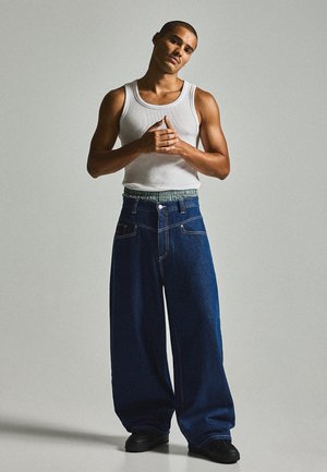 Giovane uomo con capelli corti che indossa una canotta bianca e jeans blu scuro oversize, in piedi davanti a uno sfondo chiaro e semplice.