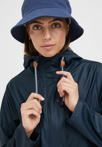 Navy waterproof raincoat with a high collar, snap buttons, and adjustable drawstrings featuring wooden accents; worn with a coordinating blue bucket hat.