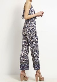 Kobieta w bezrękawnikowym kombinezonie w kwiatowy wzór z szerokimi nogawkami i beżowych platformach, stojąca na jednolitym tle.