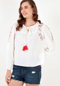 White long-sleeve blouse with red floral embroidery, a square neckline, tie detail, and red tassels. Paired with distressed denim shorts.