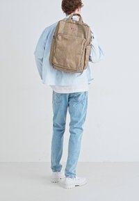 Mochila de lona beige con múltiples bolsillos, correas ajustables y un acabado texturizado. Llevada por una persona con jeans de mezclilla azul claro y zapatillas blancas.
