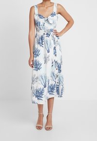 Robe midi sans manches blanche avec des motifs botaniques bleus, détail de nœud à l'avant et boutons sur le côté, portée avec des talons à brides beiges.