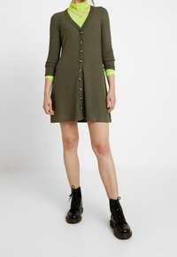 Robe côtelée vert olive à manches longues, avec boutonnière sur le devant et encolure en V. Portée sur un haut vert néon, associée à des bottes noires lacées.