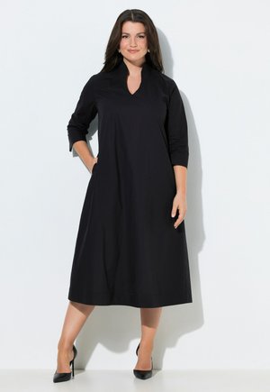 Mujer de cabello oscuro que lleva un vestido midi negro de tres cuartos de manga y tacones negros, posando contra un fondo blanco.