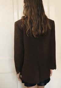 Ruskea ruudullinen bleiseri, jossa on muotoiltu leikkaus, pitkät hihat ja nappikiinnitys. Sisältää hienovaraisen tekstuurin ja kiiltävän, rakenteellisen muotoilun.