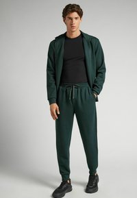 Donkergroen sportieve trainingspak met een ritsjack en broek met een koord, gemaakt van zacht materiaal, gecombineerd met zwarte sneakers.