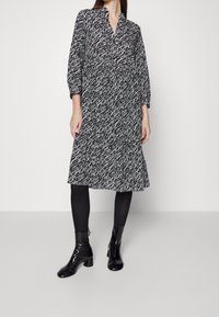 Schwarz-weiß gemustertes Kleid mit lässiger Passform, V-Ausschnitt, dreiviertel Ärmeln und ausgestelltem Rock, kombiniert mit schwarzen Knöchelstiefeln.