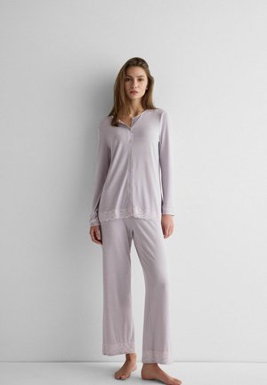 Femme debout pieds nus contre un mur, vêtue d'un ensemble de pyjama violet clair à boutons avec des bordures en dentelle aux poignets et à l'ourlet du pantalon.