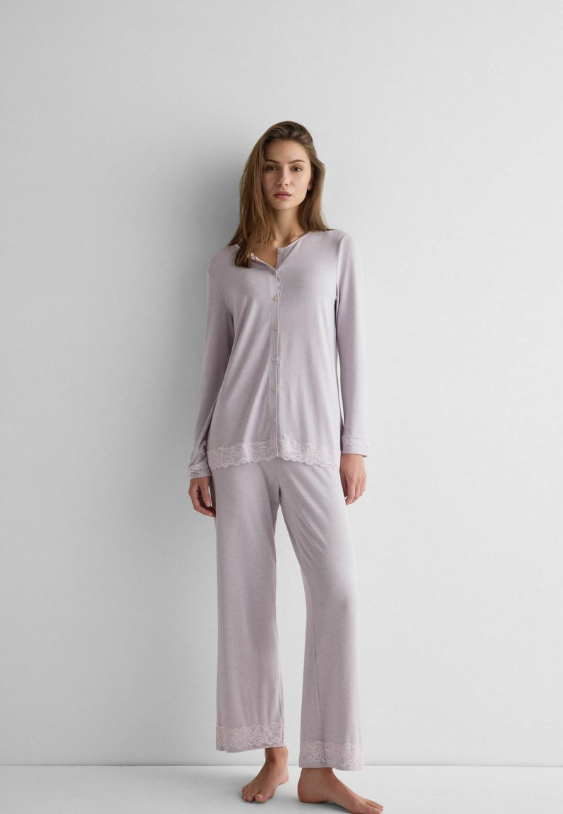 Frau steht barfuß an einer Wand und trägt einen hellvioletten, mit Knöpfen versehenen Pyjama mit Spitzensaum an den Manschetten und Hosenenden.