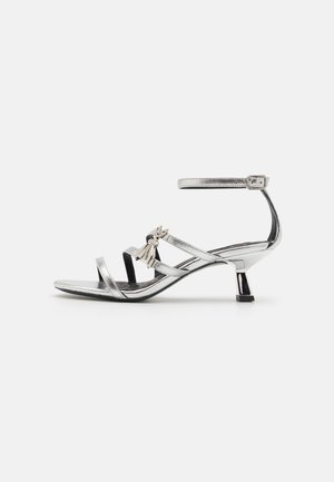 Srebrna sandala s remenčićima i niskom blok petom, koja ima dekorativni naglasak s rese i sjajnu metalik završnu obradu. Uključena je podesiva narukvica za gležanj.