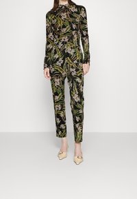Combinaison noire à manches longues présentant un motif floral vibrant en vert et blanc, avec un pantalon ajusté et un col haut. Texture de tissu lisse.