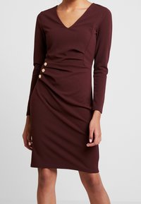 Burgundy långärmad klänning med V-hals, med draperad detaljer och tre guldfärgade knappar på sidan. Tillverkad av ett mjukt, stretchigt tyg.