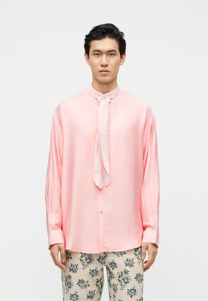 Άνδρας που φοράει μια φαρδιά ροζ σατέν μπλούζα με λεπτομέρεια δέσιμου στο λαιμό και εκρού παντελόνι με μπλε λουλουδάτο σχέδιο, στέκεται μπροστά σε λευκό φόντο.