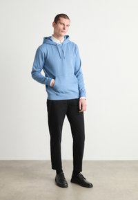 Hoodie azul claro feito de tecido macio, apresentando um bolso tipo canguru e capuz com cordão, combinado com calças pretas e sapatos pretos.